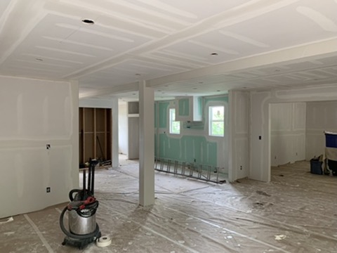The open ground floor with drywall hung and taped, a boxed beam and column carrying the span, backer board at the kitchen wall.