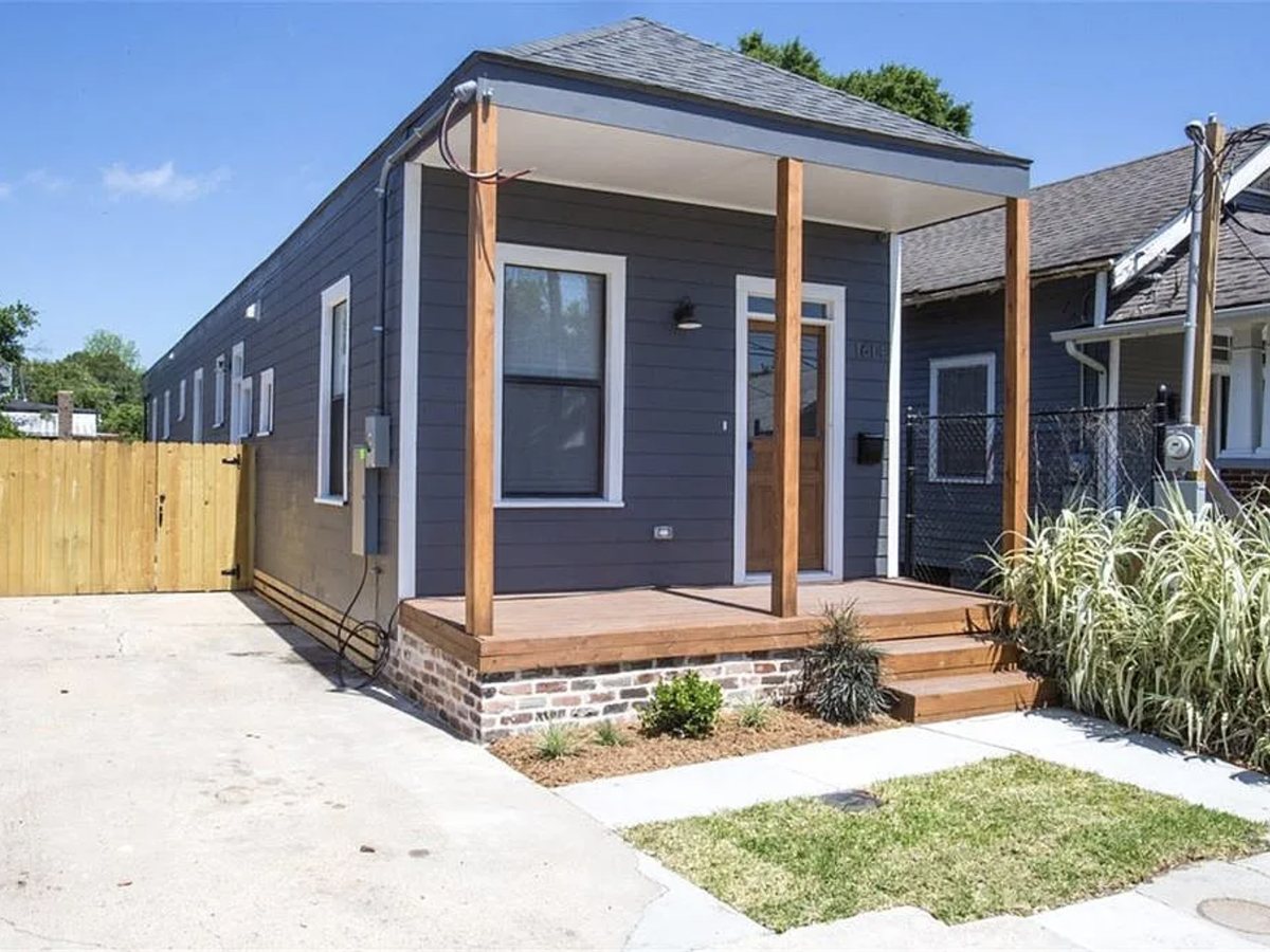 The finished house from the front corner, its full length running back along the drive, with a new porch on timber posts over a brick pier base and new siding throughout.