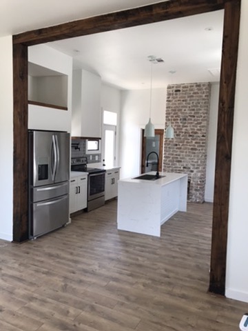 The finished kitchen seen through an opening framed in dark reclaimed timber, with the original chimney brick left exposed beside the island.