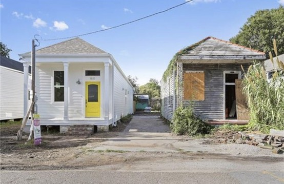 Two houses side by side on the block. On the left a completed renovation, white with a yellow front door. On the right this house, weathered gray, ivy over the roof and plywood over the openings.