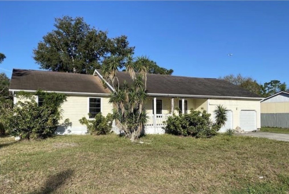 The house at purchase: a single-story ranch with a weathered shingle roof, yuccas and overgrown shrubs across the front, and a two-car garage at the right.
