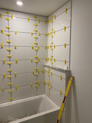 Wall tile set over the tub with yellow leveling clips still projecting from every joint.